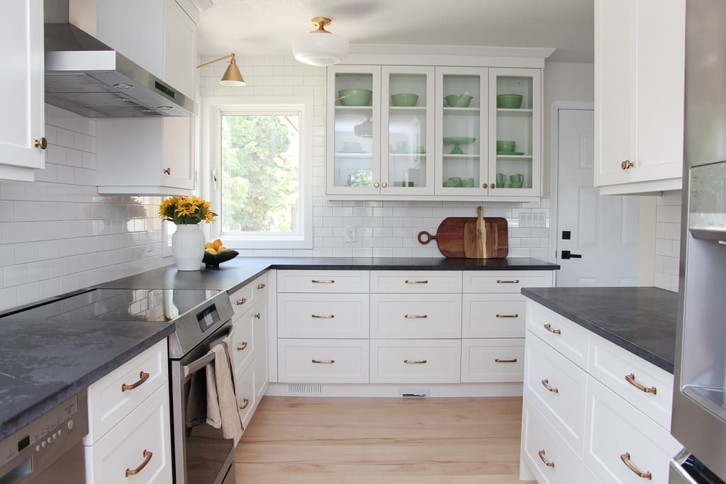 timeless white kitchen cabinets, black quartz counter, gold sconce over window, sunflowers and lemon decor, subway tile to ceiling, glass cabinets, jadeite collection