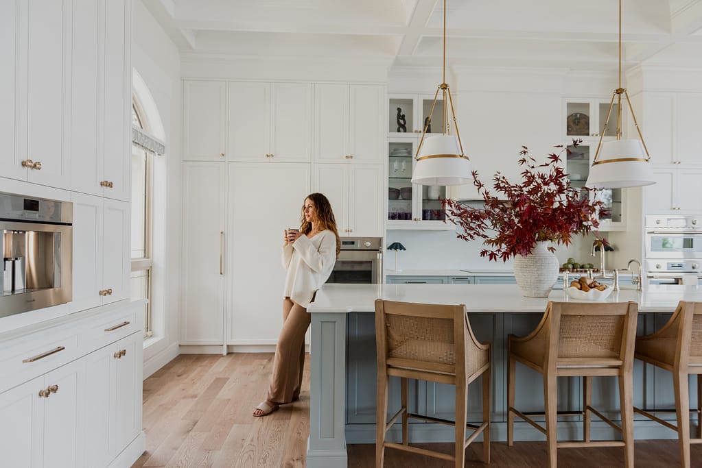 Woman in Kitchen with Range hood feature wall with integrated Thermador appliances, coffered ceilings, cane wood bar stools, and a spacious entertaining island